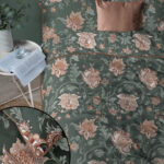 Green floral bedspread with peach flowers on a bed, plus a matching copper pillow, a potted plant, and a side table with an open book and glass of water.