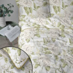 White bedding with a pale-green hydrangea and leaf pattern on a bed; inset shows close-up corner with pale yellow piping and edge detail, a side table with a plant and an open magazine nearby.
