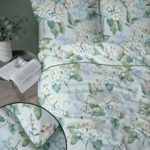 A bedroom scene with a light blue floral duvet featuring hydrangea and leafy patterns, a glass on an open book at a small table, and a potted plant nearby.